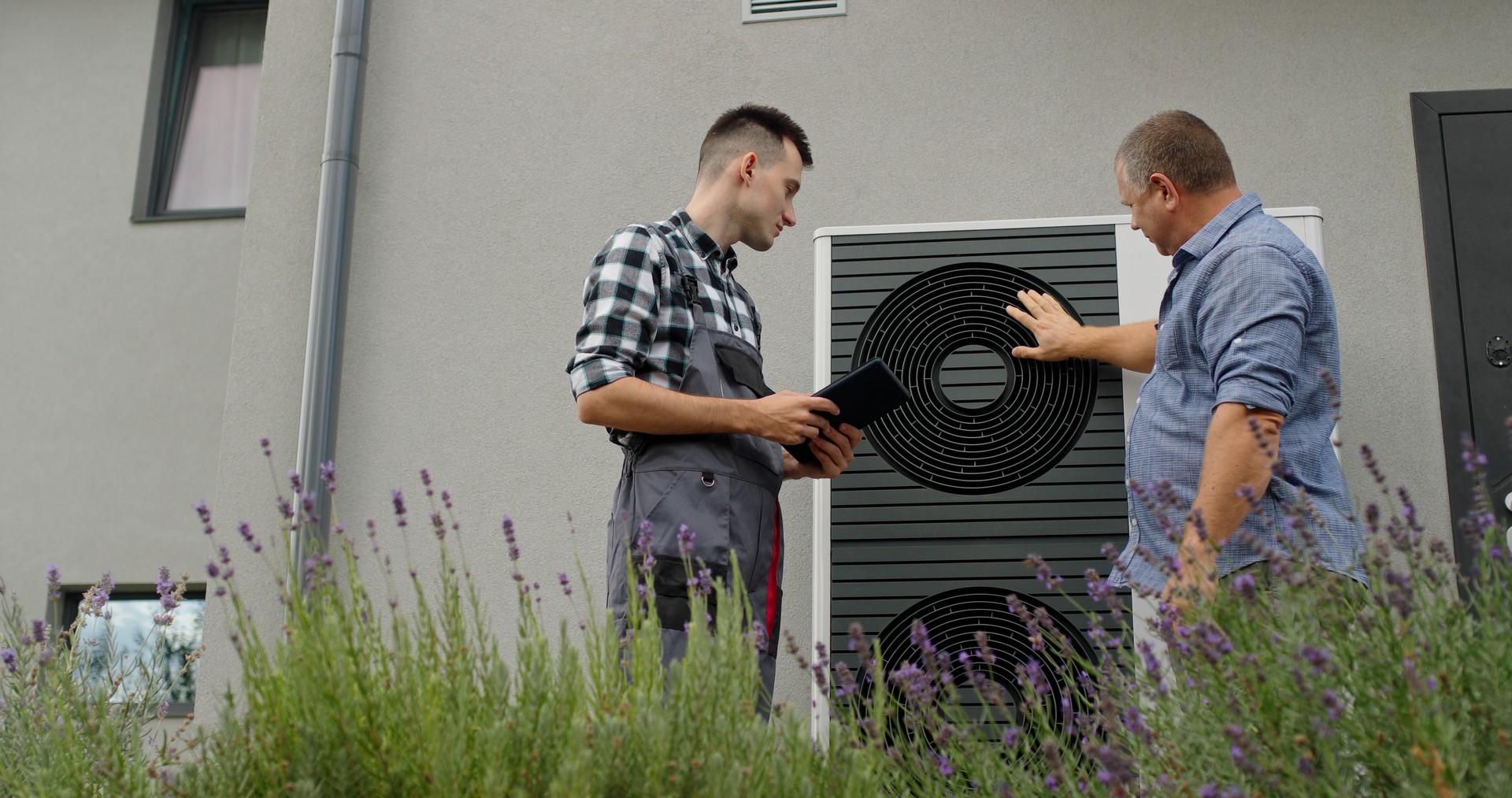 A technician and homeowner discuss heat pump maintenance, referencing a tablet near the unit.